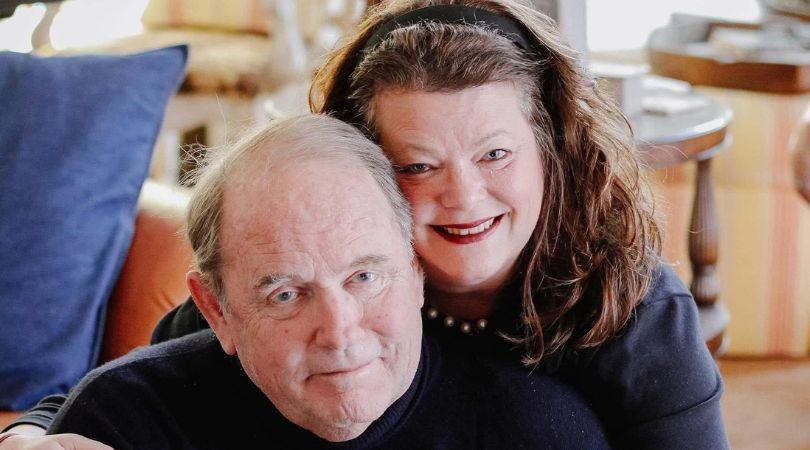 A couple embraces closely indoors; a man sits in front while a woman wraps her arms around his shoulders, smiling at the camera.